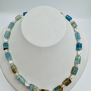 Könnte beinhalten: Eine Halskette mit rechteckigen Aquamarin-Perlen in Blau- und Brauntönen, kombiniert mit kleinen weißen Perlen. Die Perlen sind mit silberfarbenen Metallelementen verbunden, wodurch ein einzigartiges und elegantes Schmuckstück entsteht.