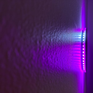 May include: A wall-mounted LED light strip emitting a vibrant purple and white glow. The light is mounted on a textured wall, creating a contrast between the smooth light and the rough surface. The LEDs are arranged in a linear fashion.