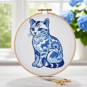 Könnte beinhalten: Eine Kreuzstich-Stickerei einer blau-weißen Katze in einem Holzreifen. Das Katzendesign weist florale Muster auf und ist vor einem weißen Hintergrund platziert. Goldene Scheren befinden sich im Vordergrund.