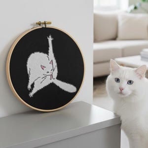 Puede incluir: Un aro de punto de cruz negro con un gato blanco con la pata levantada. El gato está sobre un fondo negro. El aro es de madera y tiene un cierre de latón.