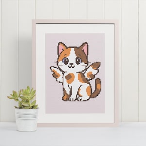 May include: A framed cross-stitch picture of a cartoon cat with brown, orange, and white markings. The cat has wings and is set against a light pink background. The frame is light-coloured, and a small potted plant sits in front.