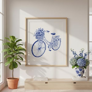 Op de afbeelding: Blauw-witte fietskunstprint in een houten lijst. De fiets heeft een mand vol bloemen. Het kunstwerk wordt tentoongesteld aan een witte muur boven een houten kast. Een potplant en een vaas met bloemen staan ook in de kamer.