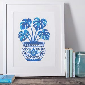 Peut inclure: Une impression encadrée représentant une broderie au point de croix bleue et blanche d'une plante Monstera en pot. La plante a de grandes feuilles fendues, et le pot est décoré de motifs floraux. L'œuvre est exposée sur une surface en bois.