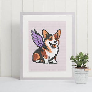 May include: Framed cross-stitch art featuring a smiling corgi with purple wings. The corgi has brown, black, and white markings. The artwork is framed in a light gray frame and set against a pale pink background. A small potted plant sits nearby.