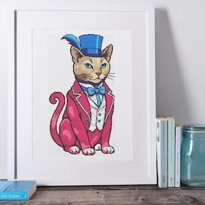 May include: Cross-stitch image of a cat in a pink suit, blue top hat with a feather, and a blue bow tie. The cat has blue eyes and a white waistcoat. The artwork is framed in a white frame and displayed on a wooden surface.
