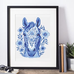 May include: A framed cross-stitch of a horse's head in shades of blue, decorated with floral motifs. The artwork is set against a white backdrop, framed in black. The piece is displayed on a wooden surface, with books and a plant nearby.