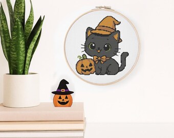 May include: A cross-stitch embroidery of a black cat wearing a witch hat and bow tie, with a jack-o'-lantern. A wooden pumpkin decoration with a witch hat sits on a stack of books. A potted plant is also in the image.
