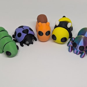 Könnte beinhalten: Eine Sammlung farbenfroher, kleiner Insektenfiguren. Die Figuren umfassen eine grüne Raupe, einen lila Marienkäfer, ein orangefarbenes Eichhörnchen, eine gelb-schwarze Biene und eine Regenbogen-Spinne. Jede Figur hat ein einzigartiges Design, vor weißem Hintergrund.