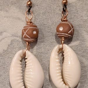May include: A pair of dangle earrings featuring white cowrie shells suspended from brown spherical beads with white etched patterns. The beads are connected to dark metal ear wires by copper-colored wire wraps, creating a cohesive design.