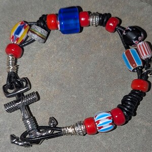 May include: A bracelet with a silver anchor charm and a variety of colourful beads. The beads include red, blue, and patterned glass elements, strung on a black leather cord. The bracelet has a heart-shaped tag that says "Made with Love."