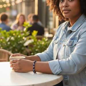 Peut inclure: Une femme porte une veste en jean bleu clair et un bracelet de perles bleues et blanches. Elle est assise à une table en marbre blanc et tient une tasse de café. L'arrière-plan est flou, avec des sièges extérieurs et de la verdure.