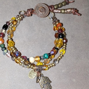 May include: A multi-strand bracelet with a mix of colorful beads in shades of yellow, orange, brown, and black. The bracelet features a bronze-colored clasp and a charm with the text "MADE WITH LOVE" and a Hamsa hand design.