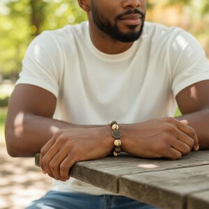 May include: A beaded bracelet with a bronze-colored centerpiece and wooden beads. The bracelet is worn on a person's wrist, resting on a wooden table. The person is wearing a white t-shirt and blue jeans.