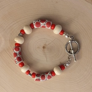 May include: A beaded bracelet featuring red and white patterned cylindrical beads, interspersed with red and off-white round beads. The bracelet has a silver-colored toggle clasp. The beads are strung on a silver-colored wire.