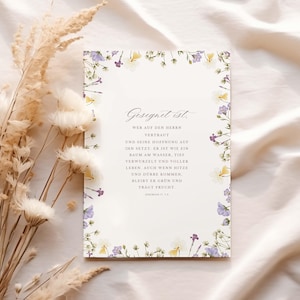 May include: A rectangular card with floral border in shades of purple, yellow, and white. The card features elegant script in German. Dried flowers and foliage are arranged to the left of the card on a cream-colored fabric.
