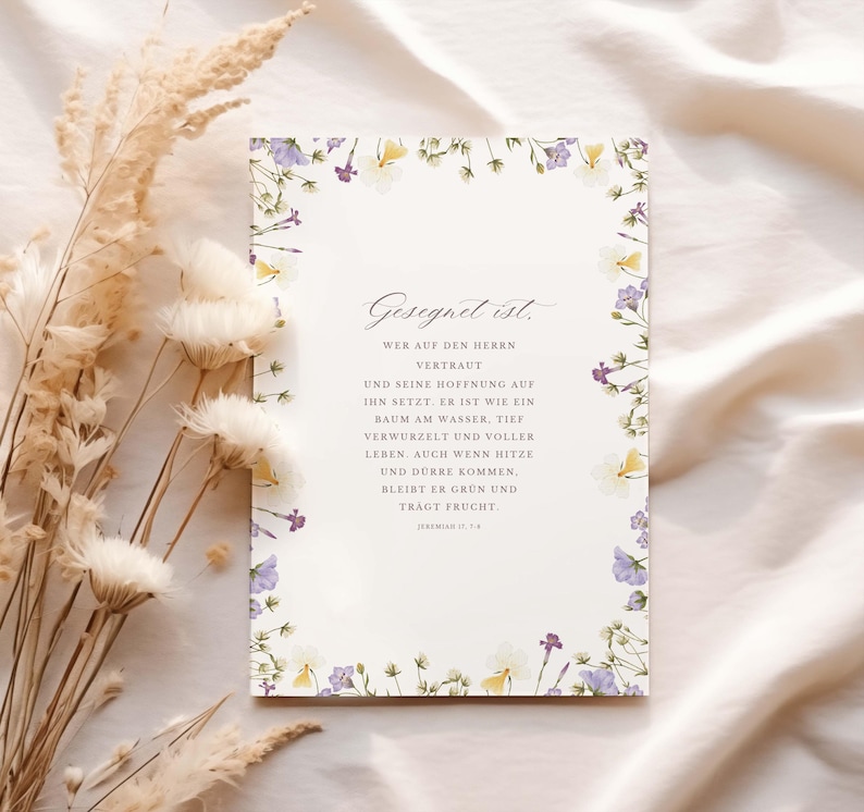 May include: A rectangular card with floral border in shades of purple, yellow, and white. The card features elegant script in German. Dried flowers and foliage are arranged to the left of the card on a cream-colored fabric.