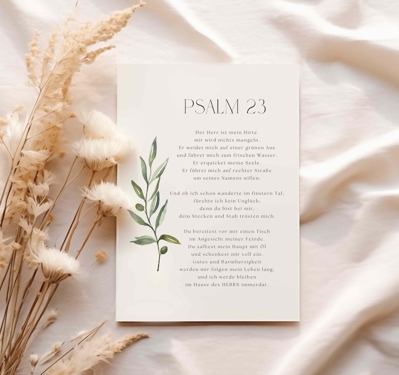 May include: A white card with the text "PSALM 23" and a German poem. The card is decorated with a watercolor olive branch and is set against a neutral background with dried flowers.