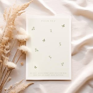 May include: A cream-colored art print with the text "PSALM 62:2" and German text. The print features small green leaf illustrations scattered across the surface. Dried flowers and foliage are in the background.