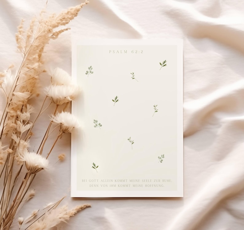 May include: A cream-colored art print with the text "PSALM 62:2" and German text. The print features small green leaf illustrations scattered across the surface. Dried flowers and foliage are in the background.