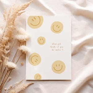 Könnte beinhalten: Eine weiße Grußkarte mit gelben Smiley-Gesichtern und dem Text "when god thinks of you, he smiles." Die Karte ist von getrockneten Blumen umgeben und liegt auf einem cremefarbenen Stoff.