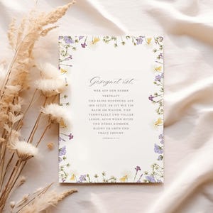 Könnte beinhalten: Eine rechteckige Karte mit floralem Rand in Lila-, Gelb- und Weißtönen. Die Karte zeigt elegante Schrift in deutscher Sprache. Getrocknete Blumen und Blätter sind links von der Karte angeordnet, vor einem cremefarbenen Stoffhintergrund.