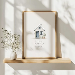 Könnte beinhalten: Ein gerahmter Kunstdruck mit einer Aquarellillustration eines weißen Hauses mit blauer Tür und Topfpflanzen. Der Druck enthält deutschen Text und wird auf einem Holzregal mit einer kleinen Vase und Grünzeug präsentiert.