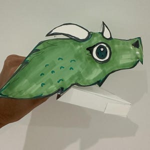 Puede incluir: Un títere de dragón de papel verde con un cuerno blanco y un gran ojo azul. El títere está sostenido por una mano.