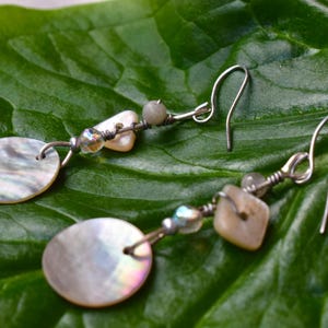 May include: Handmade earrings with iridescent, circular mother-of-pearl pendants. The earrings have silver-coloured hooks and are decorated with small, irregularly-shaped beads and square beads. The earrings are displayed on a large, green leaf.
