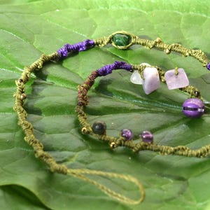 May include: Two handmade macrame bracelets. The bracelets are olive green with purple accents and feature various gemstones, including amethyst and a green stone. The bracelets are displayed on a large green leaf.