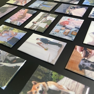 May include: A collection of square photo magnets featuring various images, including a wedding couple, a baby, a dog, and a group of people. The magnets are arranged in a grid pattern on a black surface.