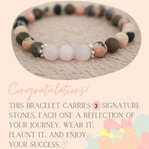 May include: A pink and brown bracelet with three larger pink stones. The bracelet has the text "Congratulations! This bracelet carries 3 signature stones, each one a reflection of your journey. Wear it, flaunt it, and enjoy your success."