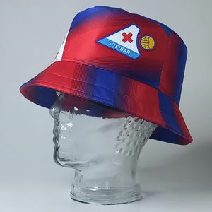 May include: A red, white, and blue bucket hat with a geometric pattern. The hat features a patch with a red cross, a soccer ball, and the text "SDEIBAR". The hat is displayed on a clear glass head.