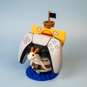 Może przedstawiać: Biało-czarny kontroler PlayStation 5 spoczywający na niestandardowym stojaku. Stojak ma żółty design statku z piracką flagą, postacią z kreskówki i niebieską podstawą. Unikalne akcesorium do gier.