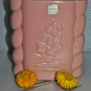 May include: A pink ceramic planter with a raised design of a ship on the front. The planter has a label that reads "Handmade, U.S.A."