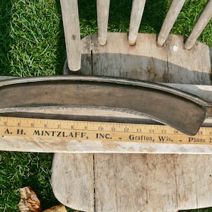 May include: A vintage metal scythe blade, curved with a hooked end, lies on a wooden surface with a ruler. The ruler shows the text "A. H. Mintzlaff, Inc."