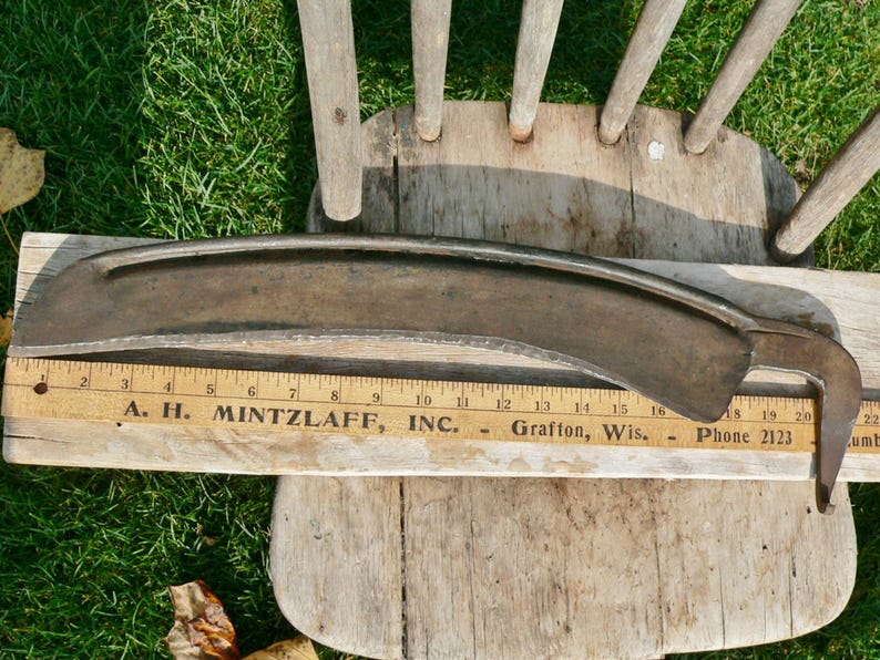 May include: A vintage metal scythe blade, curved with a hooked end, lies on a wooden surface with a ruler. The ruler shows the text "A. H. Mintzlaff, Inc."