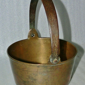 May include: A vintage brass pot with a rounded body and a sturdy handle. The pot has a worn, aged patina and is perfect for rustic decor.