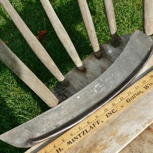 May include: A weathered metal scythe blade with a curved edge, alongside a wooden ruler. The ruler displays the text "H. MINTZLAFF, INC.". The scythe is near a wooden chair. The blade's length is approximately 30 cm.