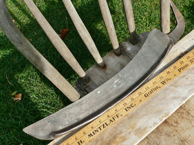 May include: A weathered metal scythe blade with a curved edge, alongside a wooden ruler. The ruler displays the text "H. MINTZLAFF, INC.". The scythe is near a wooden chair. The blade's length is approximately 30 cm.