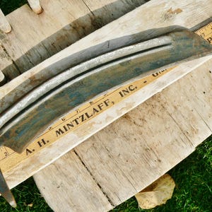 May include: A vintage, curved metal blade with a weathered, teal and silver finish. The blade rests on a wooden surface with a vintage ruler that reads "A. H. MINTZLAFF, INC." and "Phone 2123". The blade's design suggests it may be a scythe or similar tool.