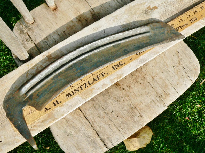 May include: A vintage, curved metal blade with a weathered, teal and silver finish. The blade rests on a wooden surface with a vintage ruler that reads "A. H. MINTZLAFF, INC." and "Phone 2123". The blade's design suggests it may be a scythe or similar tool.