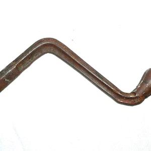May include: A rusty, bent metal crank handle with a rounded grip. The handle is a dark brown color and appears to be made of iron or steel. The handle has a hexagonal end and a curved design.