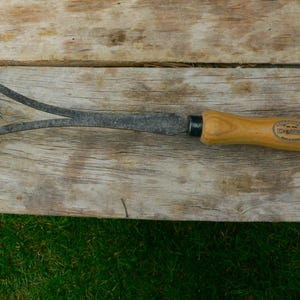May include: A hand-held garden cultivator featuring a dark, forged metal head with four curved tines and a light-colored wooden handle. The handle displays a black oval brand logo with the text DeWit and Made in Holland, and includes a circular hole.