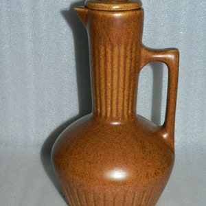 May include: A brown ceramic pitcher with a lid and a handle. The pitcher has a textured surface and a ribbed design.