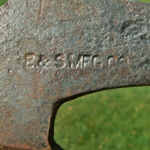 May include: Close-up of a weathered, rusty metal tool with the embossed text "E & SMFG Co." The tool has a curved shape with a large opening. The background is a blurred green.