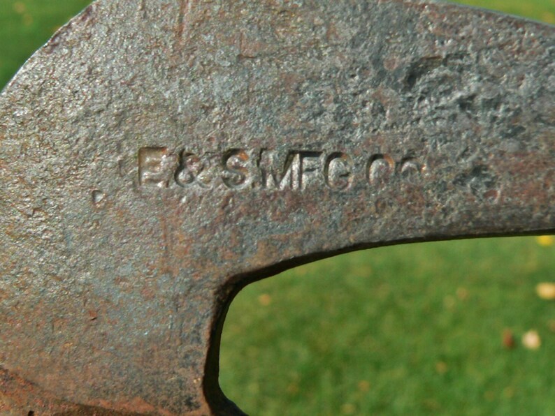 May include: Close-up of a weathered, rusty metal tool with the embossed text "E & SMFG Co." The tool has a curved shape with a large opening. The background is a blurred green.