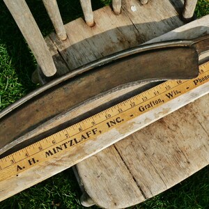 May include: An antique, curved metal scythe blade with a wooden ruler measuring in inches. The blade is a dark, aged metal, and the ruler displays the text "A. H. MINTZLAFF, INC." and "Grafton, Wis."