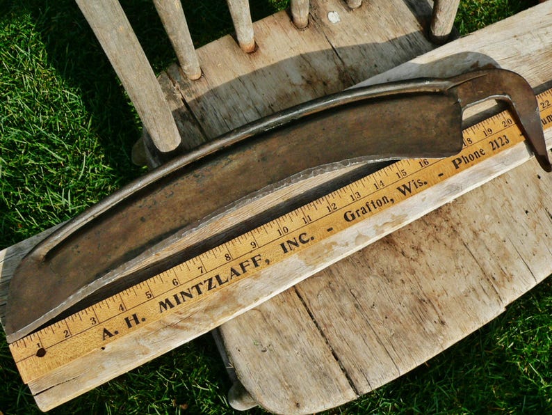 May include: An antique, curved metal scythe blade with a wooden ruler measuring in inches. The blade is a dark, aged metal, and the ruler displays the text "A. H. MINTZLAFF, INC." and "Grafton, Wis."
