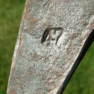 May include: Close-up of a weathered, metallic object with a rough, textured surface. The object has a dark, aged appearance with a stamped marking of "47". The background is a blurred green.