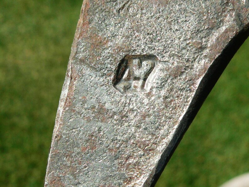 May include: Close-up of a weathered, metallic object with a rough, textured surface. The object has a dark, aged appearance with a stamped marking of "47". The background is a blurred green.